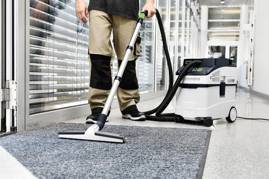 Festool CLEANTEC CT 25 Absaugmobil reinigt Teppich mit Bodendüse, Absauganlage im Hintergrund sichtbar.