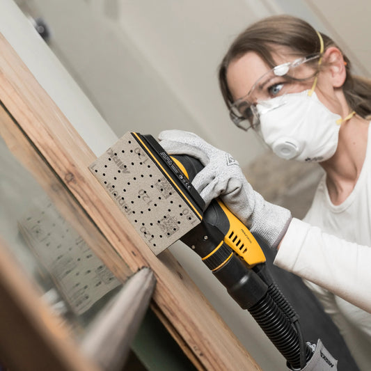 Frau schleift mit Mirka Schleifmaschine und Iridium Schleifpapier ein Holzfenster – präzises, staubfreies Arbeiten mit Absaugung und Schutzmaske.